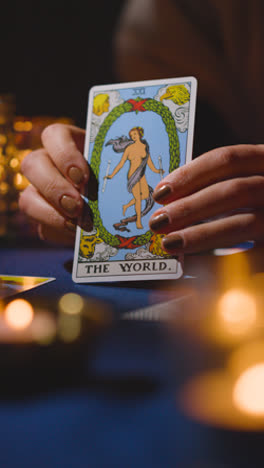 Video-Vertical-De-Cerca-De-Una-Mujer-Dando-Lectura-De-Cartas-Del-Tarot-En-Una-Mesa-A-La-Luz-De-Las-Velas-Sosteniendo-La-Carta-Mundial